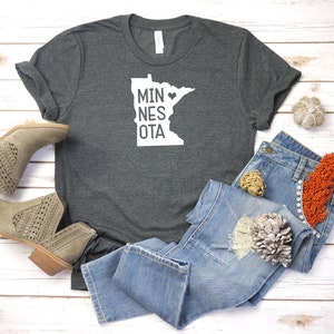 May include: A dark gray t-shirt with a white outline of the state of Minnesota and the text "MINNESOTA" with a heart in place of the "O". The shirt is laid out on a white wooden surface with a pair of brown ankle boots, blue jeans, and fall-themed decorations.