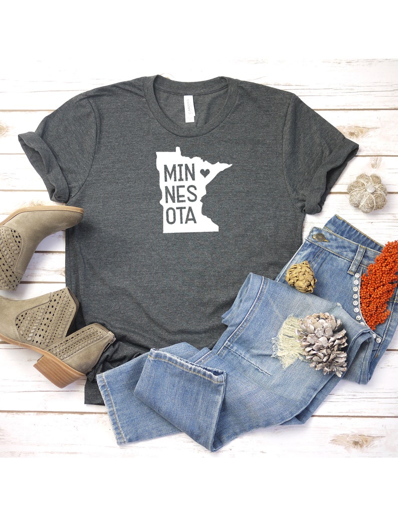 May include: A dark gray t-shirt with a white outline of the state of Minnesota and the text "MINNESOTA" with a heart in place of the "O". The shirt is laid out on a white wooden surface with a pair of brown ankle boots, blue jeans, and fall-themed decorations.