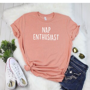 May include: A peach-colored t-shirt with the text "NAP ENTHUSIAST" printed in white. The shirt is laid out on a white fuzzy surface with a pair of white sneakers and a pair of blue denim shorts with rhinestones.