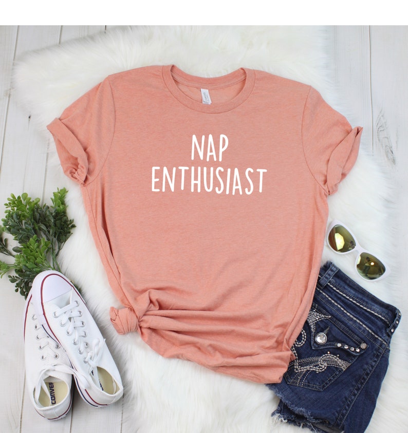 May include: A peach-colored t-shirt with the text "NAP ENTHUSIAST" printed in white. The shirt is laid out on a white fuzzy surface with a pair of white sneakers and a pair of blue denim shorts with rhinestones.