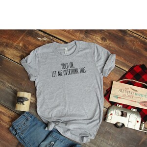 May include: A gray t-shirt with the text "HOLD ON, LET ME OVERTHINK THIS" printed in black. The shirt is laid flat on a wooden surface with a pair of blue jeans, a small wooden sign, and a toy camper.