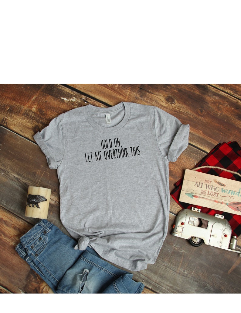 May include: A gray t-shirt with the text "HOLD ON, LET ME OVERTHINK THIS" printed in black. The shirt is laid flat on a wooden surface with a pair of blue jeans, a small wooden sign, and a toy camper.