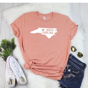 May include: A peach-colored t-shirt with a white outline of the state of North Carolina and the text "I love North Carolina" with a heart replacing the "I".