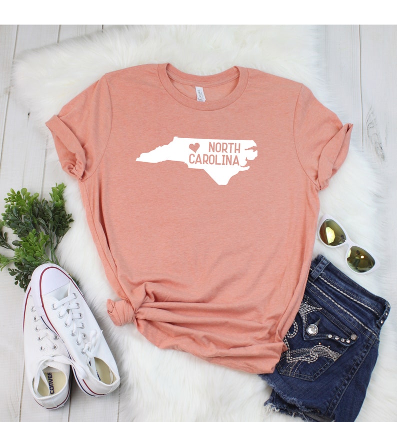 May include: A peach-colored t-shirt with a white outline of the state of North Carolina and the text "I love North Carolina" with a heart replacing the "I".