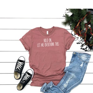 May include: A dusty rose t-shirt with the text "Hold on, let me overthink this" in white. The t-shirt is paired with blue jeans and black sneakers.