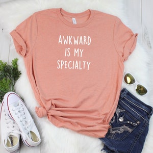 May include: A peach-colored t-shirt with the text "AWKWARD IS MY SPECIALTY" printed in white.