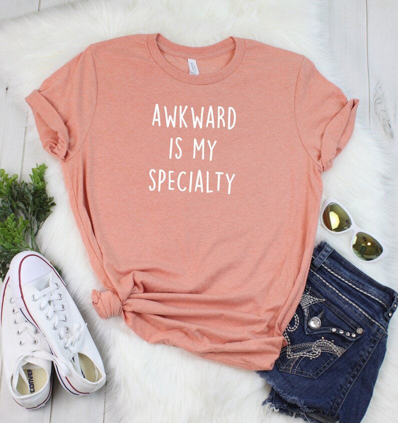 May include: A peach-colored t-shirt with the text "AWKWARD IS MY SPECIALTY" printed in white.