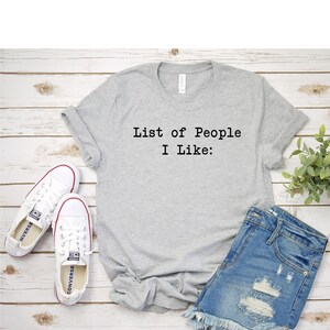 May include: A gray t-shirt with the text "List of People I Like:" printed on the front. The shirt is paired with denim shorts and white sneakers.