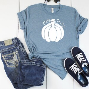May include: A light blue t-shirt with a white pumpkin graphic. The shirt is paired with blue jeans and navy blue sneakers.