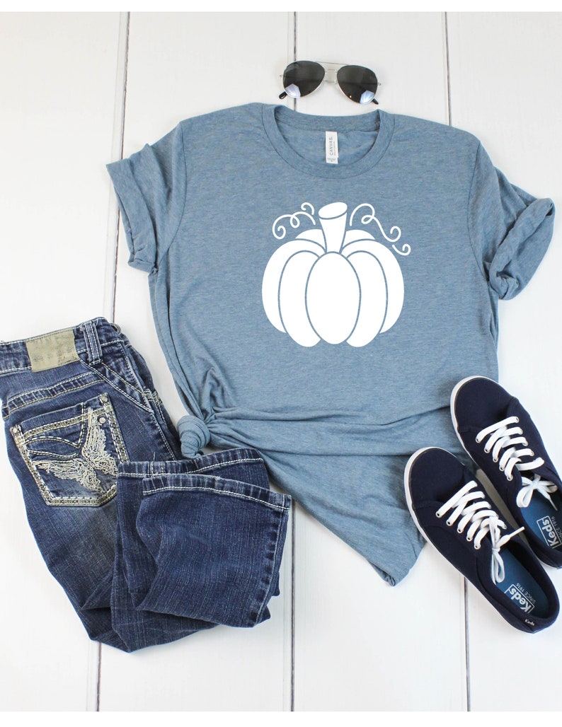 May include: A light blue t-shirt with a white pumpkin graphic. The shirt is paired with blue jeans and navy blue sneakers.