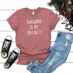 May include: A mauve t-shirt with the text "AWKWARD IS MY SPECIALTY" in white lettering. The t-shirt is paired with blue jeans and black sneakers.