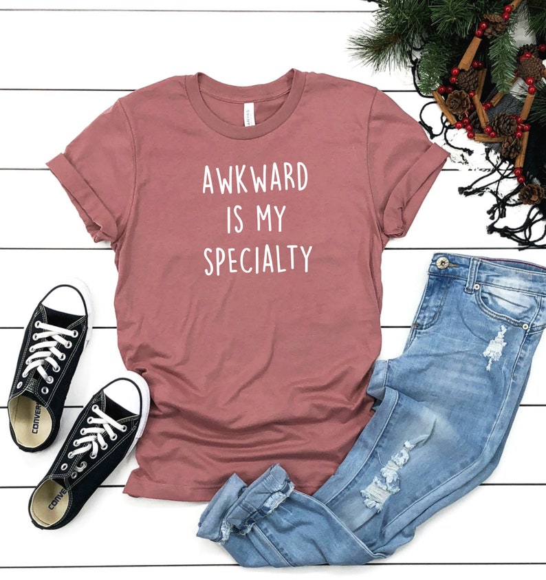 May include: A mauve t-shirt with the text "AWKWARD IS MY SPECIALTY" in white lettering. The t-shirt is paired with blue jeans and black sneakers.