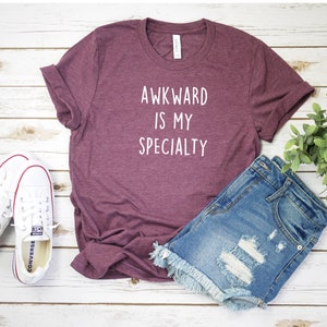 May include: A maroon t-shirt with the text "Awkward is my specialty" in white lettering.