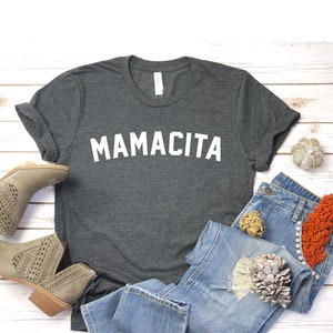 Pu&ograve; includere: Una maglietta grigia scura con la parola "MAMACITA" stampata in bianco sul davanti. La maglietta &egrave; appoggiata su una superficie di legno bianco con un paio di stivaletti marroni chiari, jeans blu e oggetti decorativi.