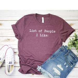 May include: A maroon t-shirt with the text "List of People I Like:" printed in white on the front. The t-shirt is laying on a white wooden surface with a pair of white sneakers and a pair of blue denim shorts.
