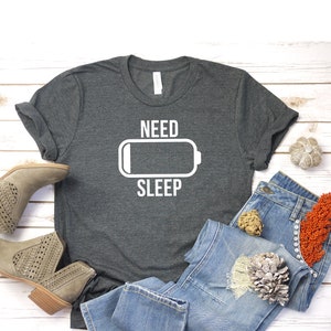 May include: A dark gray t-shirt with a white graphic of a battery icon and the text "NEED SLEEP". The shirt is laid out on a white wooden surface with a pair of brown ankle boots, blue jeans, and decorative items.