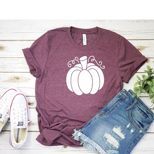 May include: A maroon t-shirt with a white pumpkin graphic. The shirt is laying on a white wooden surface with a pair of white sneakers and denim shorts.