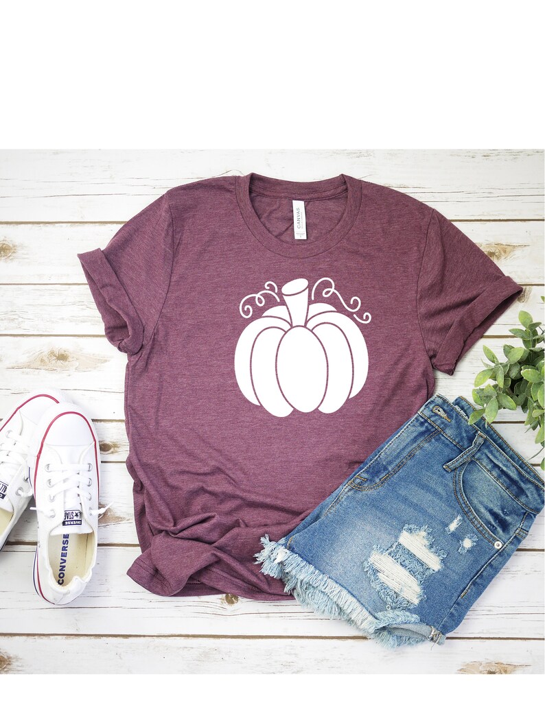 May include: A maroon t-shirt with a white pumpkin graphic. The shirt is laying on a white wooden surface with a pair of white sneakers and denim shorts.