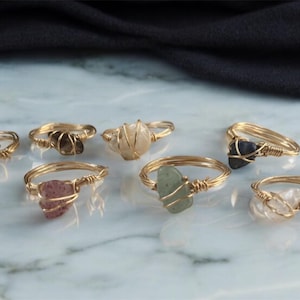 Puede incluir: Siete anillos de oro con piedras de diferentes colores. Cada anillo tiene un alambre envuelto alrededor de la piedra. Las piedras son verdes, marrones, blancas, rosas y azules.