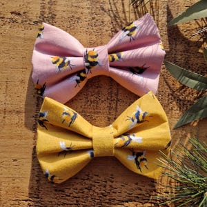 May include: Two fabric bow ties with a bee pattern. One bow tie is pink with black bees and the other is yellow with black bees.