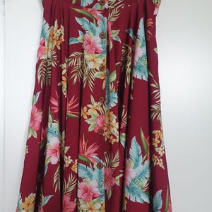 May include: A burgundy button-down skirt with a vibrant floral pattern. The skirt features a tropical design with pink, yellow, and blue flowers and green leaves. The skirt is hanging on a wooden hanger.