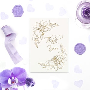 Pode incluir: Um cart&atilde;o branco com um design floral dourado e o texto "Obrigado". O cart&atilde;o &eacute; cercado por cora&ccedil;&otilde;es roxos e uma fita roxa.