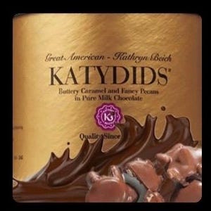 May include: A gold-colored container of KATYDIDS candy. The label reads "Great American - Kathryn Beich" and describes the product as "Buttery Caramel and Fancy Pecans in Pure Milk Chocolate." A splash of chocolate and pecan pieces are visible.
