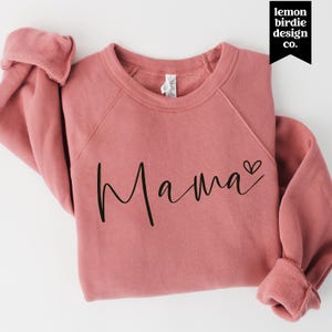 Peut inclure: Un sweat-shirt à col rond de couleur rose poudré avec le mot Mama écrit en écriture cursive noire. Le texte est placé au centre de la poitrine et comprend une petite icône de cœur noir à la fin du mot.