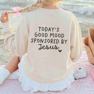 May include: Cream-colored sweatshirt with the text "TODAY'S GOOD MOOD SPONSORED BY Jesus" in black script. The sweatshirt has a crew neck and long sleeves. The model is wearing a white skirt with a ruffled hem.