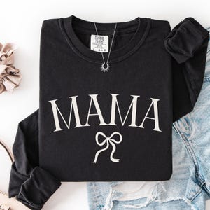 May include: Black long-sleeved sweatshirt featuring the word "MAMA" in white, decorative font, and a white bow graphic. A silver necklace with a sunburst pendant is visible. The sweatshirt is folded with denim jeans.