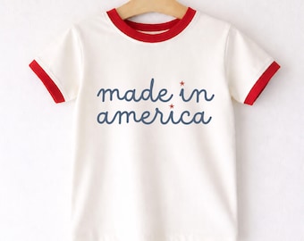 May include: Cream-colored t-shirt with red trim on the collar and sleeves. The words "made in america" are written in blue script across the front, with small red star accents. The shirt is hanging on a wooden hanger.