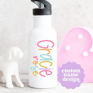 May include: White water bottle with a black cap, featuring the name "Gracie Rose" in colorful, playful lettering. A light purple sticker with the text "custom name design" is visible. A white dog figurine and a pink heart-shaped light are in the background.