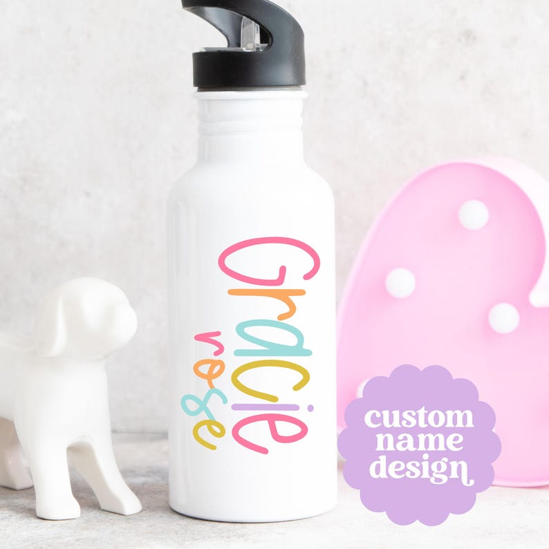 May include: White water bottle with a black cap, featuring the name "Gracie Rose" in colorful, playful lettering. A light purple sticker with the text "custom name design" is visible. A white dog figurine and a pink heart-shaped light are in the background.
