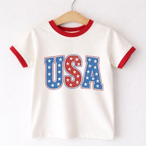 May include: A white t-shirt with red trim on the collar and sleeves. The word "USA" is printed on the front in red and blue, with a star pattern inside each letter. The shirt is hanging on a wooden hanger.