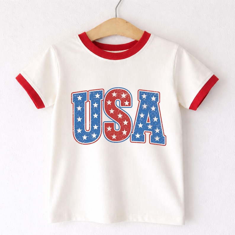May include: A white t-shirt with red trim on the collar and sleeves. The word "USA" is printed on the front in red and blue, with a star pattern inside each letter. The shirt is hanging on a wooden hanger.