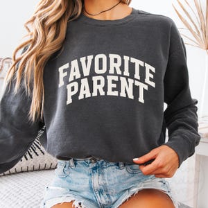 May include: Dark gray sweatshirt with the text "FAVORITE PARENT" in bold white letters. The sweatshirt has a crew neck and long sleeves. The image shows a casual style, suitable for everyday wear.