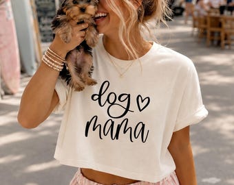 May include: Cream-coloured crop top with the text "dog ♡ mama" in black script. The woman is holding a small dog with a pink bow. She is wearing bracelets and a necklace. The background is blurred.