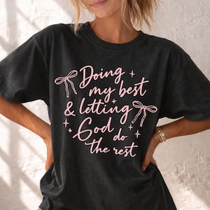 May include: Dark gray t-shirt with the text "Doing my best & letting God do the rest" in pink script. The design includes bow accents and star shapes. The shirt has short sleeves.