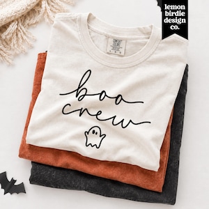May include: A cream-colored short-sleeve t-shirt with a black graphic print that reads "boo crew" in a cursive script font above a small ghost illustration. The shirt features a Comfort Colors brand label on the inside of the collar.
