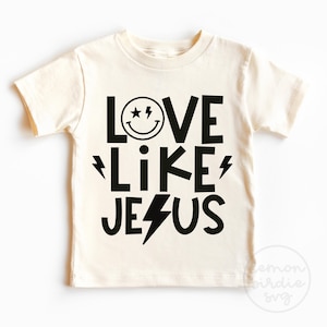 Peut inclure: Un t-shirt blanc avec du texte noir qui dit "Love Like Jesus" avec un visage souriant et des éclairs.