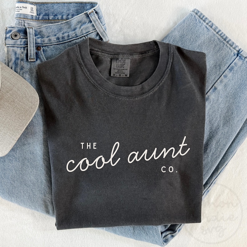 May include: A charcoal gray t-shirt with the text "THE cool aunt CO." in white script. The shirt is folded and placed on top of a pair of light blue denim jeans and a gray baseball cap.