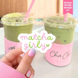 May include: A die-cut sticker featuring the text "matcha girly" in a playful, rounded font with alternating pink and green letters, accented by a small green heart. The sticker is held in front of two clear plastic cups filled with iced matcha.