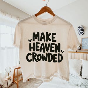 May include: A cream-coloured, short-sleeved t-shirt with the phrase "MAKE HEAVEN CROWDED" printed in bold, black letters. The shirt is hanging on a wooden hanger.