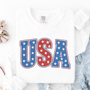 May include: White long-sleeve shirt with the word "USA" in red and blue, star-filled letters. The shirt has a distressed, vintage look. The tag on the shirt reads "Comfort Colors".