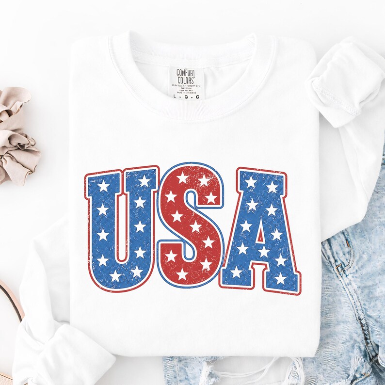 May include: White long-sleeve shirt with the word "USA" in red and blue, star-filled letters. The shirt has a distressed, vintage look. The tag on the shirt reads "Comfort Colors".