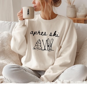 May include: Cream-colored sweatshirt with the words "apres ski" in black script. Below the text is a graphic of a pine tree and skis. A person is holding a beige mug with a black tree design.