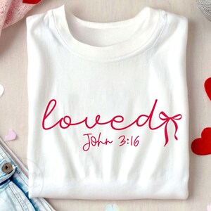 May include: White t-shirt with the word "loved" written in pink cursive script with a pink bow. The text "John 3:16" is written in pink below the word "loved".