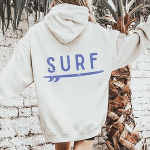 Puede incluir: Una sudadera con capucha de color crema con un gráfico azul de una tabla de surf y la palabra "SURF" en la espalda.