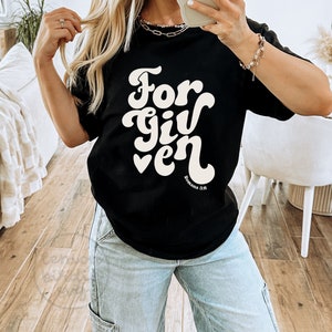 May include: Black t-shirt with white text that reads "Forgiven" in a retro font. The text is accompanied by a small heart and the words "Romans 5:8".