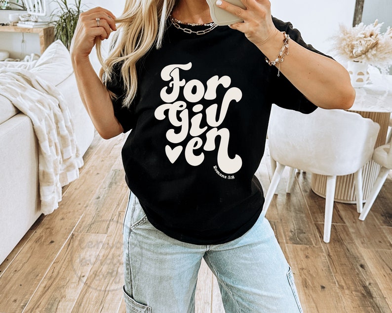 May include: Black t-shirt with white text that reads "Forgiven" in a retro font. The text is accompanied by a small heart and the words "Romans 5:8".
